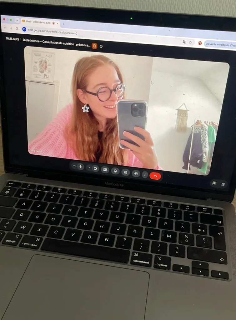 Une jeune femme aux lunettes et aux cheveux roux, souriante, participe à une consultation de nutrition en visioconférence sur un MacBook Air.