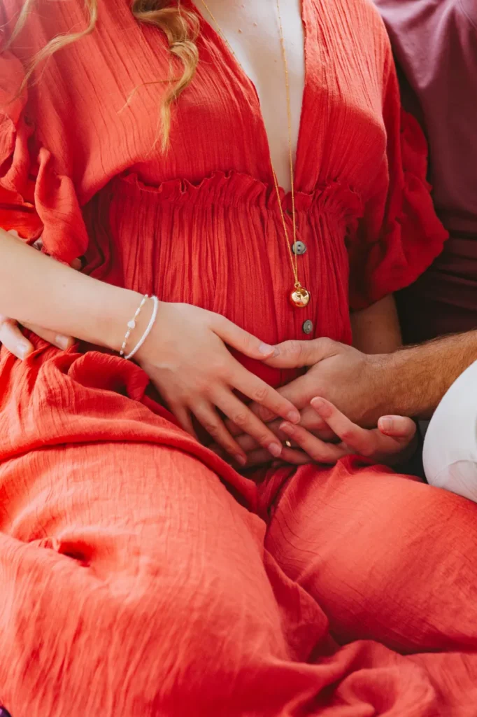 Un couple joint ses mains sur le ventre arrondi d'une femme enceinte, vêtue d'une robe rouge fluide, portant un collier bola de grossesse.