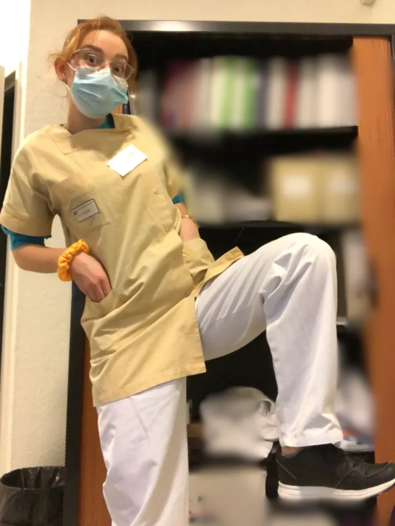 Jeune femme portant une tenue d'étudiant infirmier, masque bleu, lunettes et chaussette orange au poignet, pose près d'une étagère.