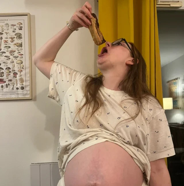 Une femme enceinte, vêtue d'un pyjama à motifs, s'apprête à manger un éclair au chocolat. Son ventre est bien rond.
