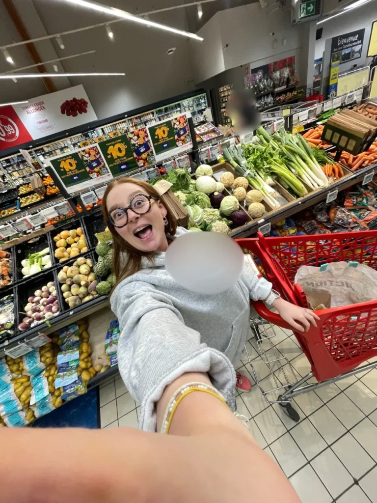 Une jeune femme souriante avec des lunettes fantaisie se prend en selfie dans un supermarché, poussant un caddie rouge rempli de légumes.