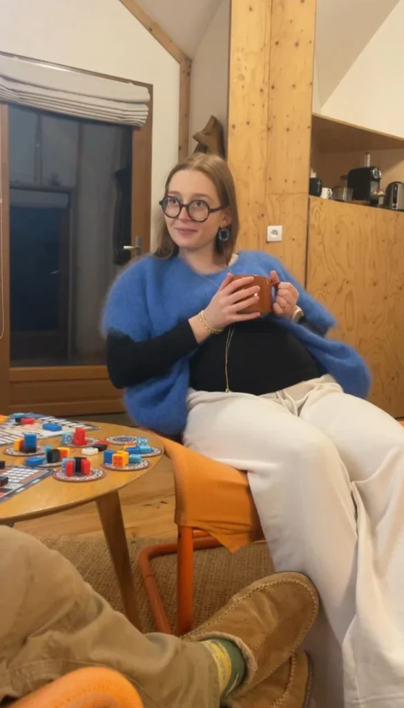Une jeune femme aux lunettes rondes, vêtue d'un gilet bleu et d'un haut noir, tient une tasse et sourit, assise près d'une table de jeux.