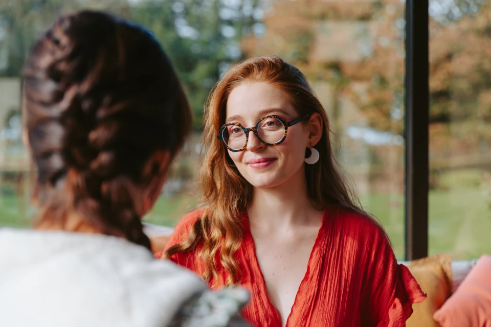 Une femme aux cheveux roux et aux lunettes rondes sourit en discutant avec une personne de dos