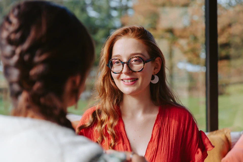 Une femme rousse souriante avec des lunettes rondes et une robe rouge discute avec une autre personne dont on ne voit que le dos.