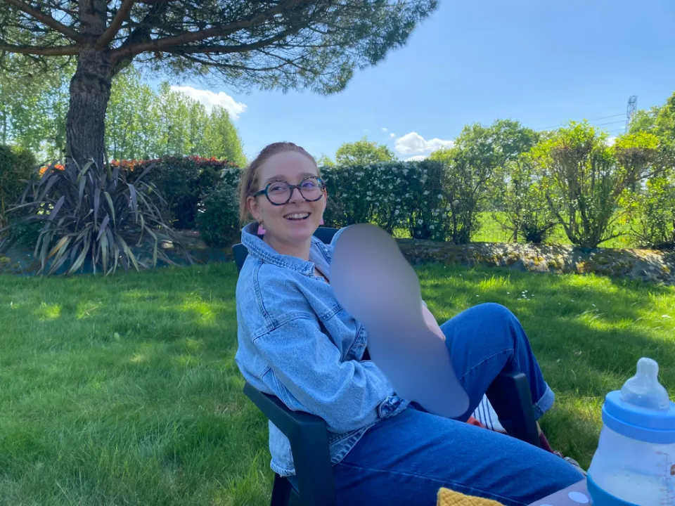 Une jeune femme avec des lunettes noires et des boucles d'oreilles roses sourit à la caméra, assise dans un jardin verdoyant par une journée ensoleillée. Elle porte une veste en jean et un jean.