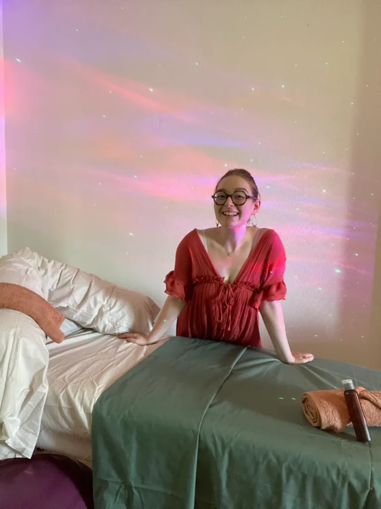 Une femme souriante avec des lunettes, portant une robe rouge, est assise au bord d'une table de massage dans un cabinet aux lumières douces et colorées.