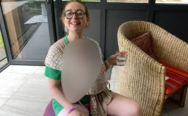 Femme souriante assise sur un ballon de fitness violet dans une véranda, tenant un verre d'eau.
