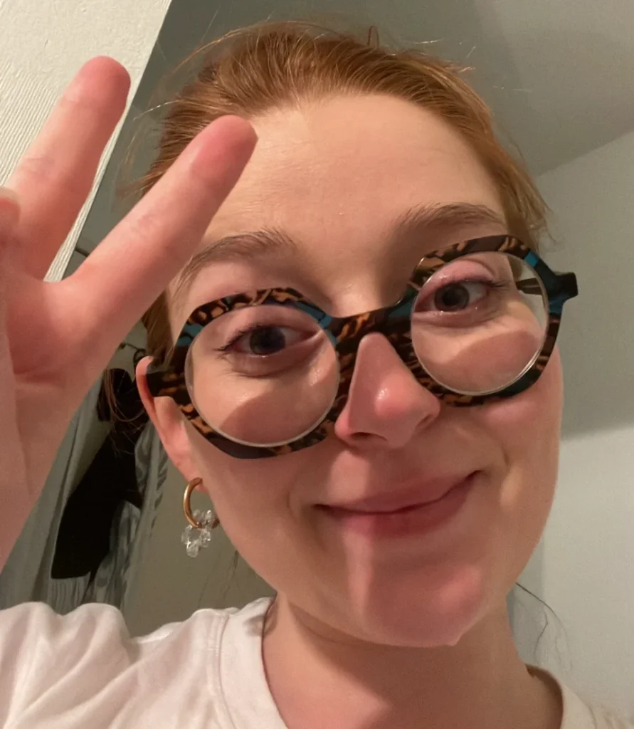 Jeune femme aux cheveux roux, portant des lunettes originales et un t-shirt blanc, fait le signe de paix avec ses doigts.