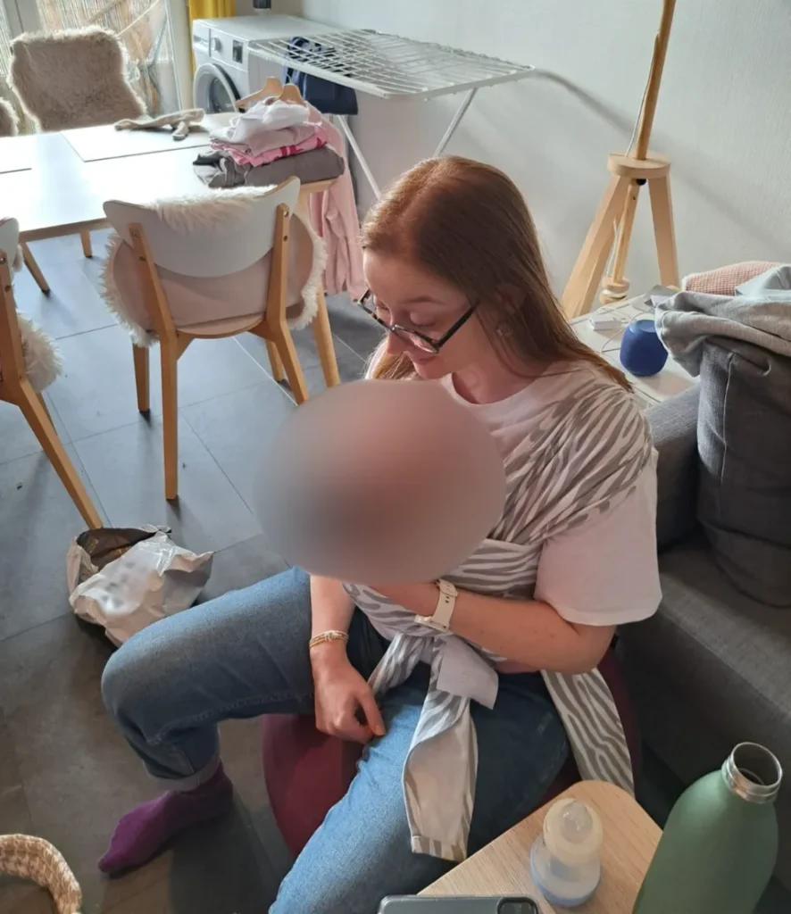 Femme rousse aux lunettes tenant un bébé dans une écharpe de portage, assise sur un ballon de yoga.