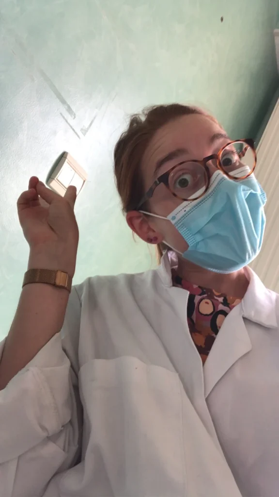 Médecin portant un masque bleu, des lunettes rondes et une blouse blanche, tenant un petit objet blanc dans sa main droite.