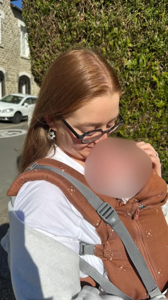 Femme avec des lunettes tenant un bébé dans un porte-bébé brun à l'extérieur, près d'un mur de pierres et d'arbustes.